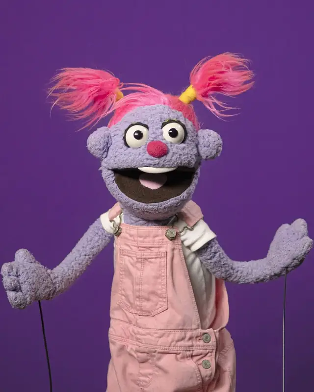 Pitzy, títere morada con coletas rosas y overol