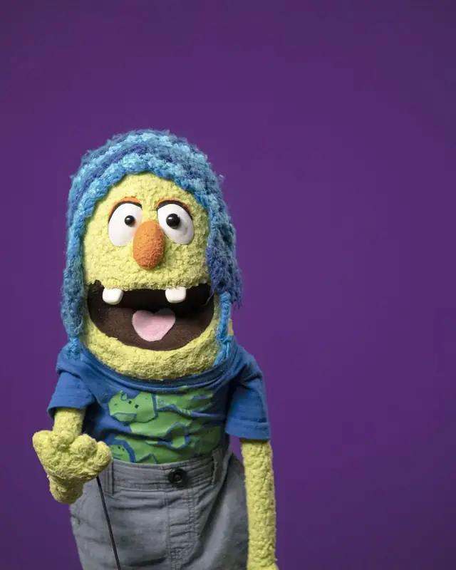 Wizz, títere verde con gorro de estambre azul y camiseta de dinosaurios